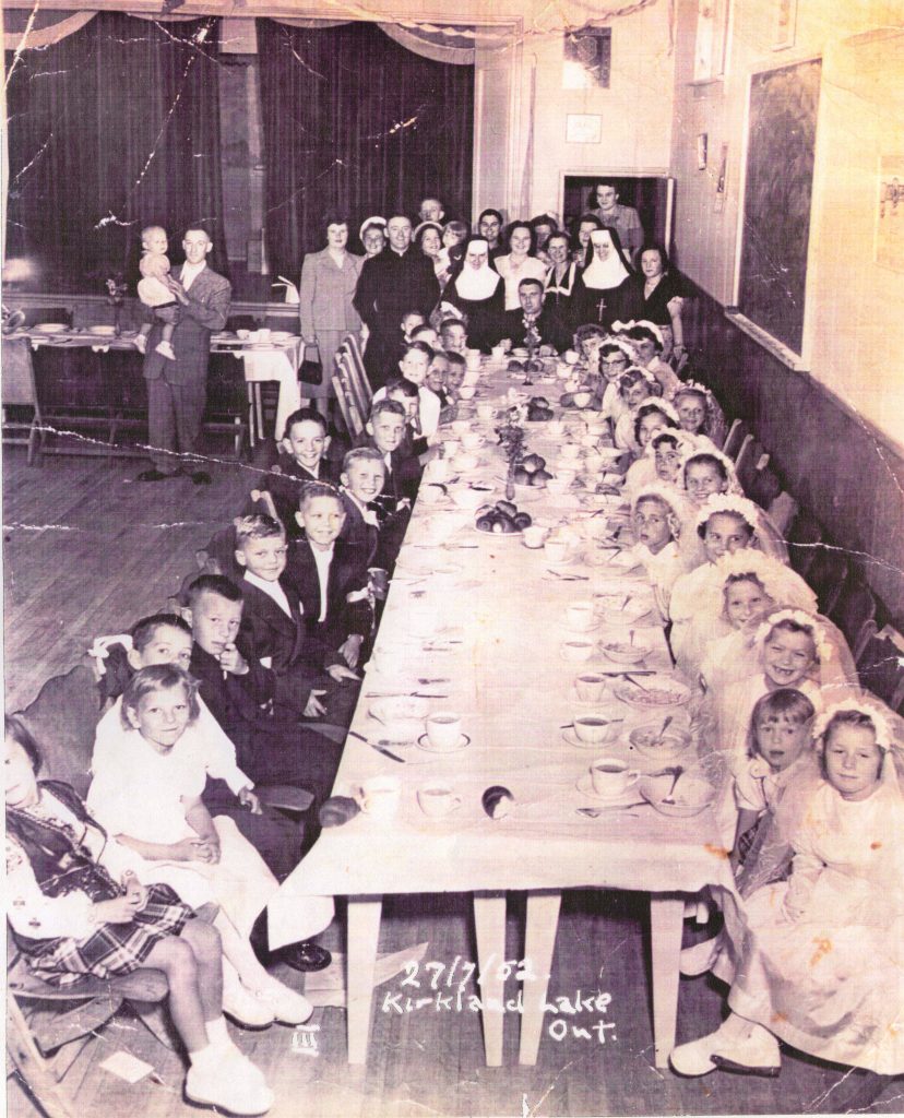 Black and white photograph of about 40 children sitting around a long table having a meal. Boys are dressed in dark suits, girls in mostly white dresses. Standing in the background of the hall are men and women, with a priest and two nuns.