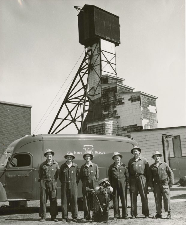 Black and white photograph of six men in overalls with their equipment in front of them. They are standing in front of their van. A mine headframe is in the background.
