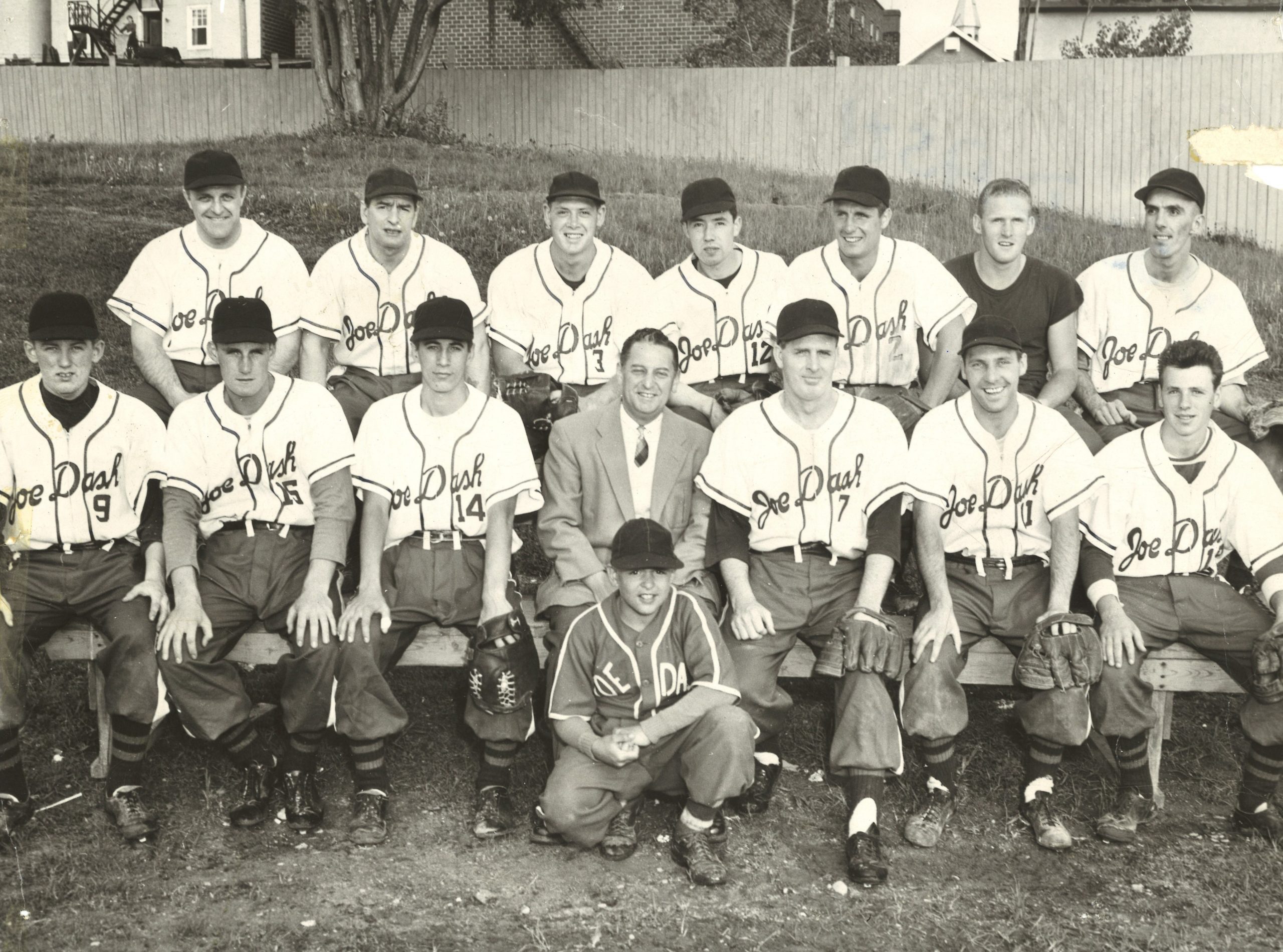Black and white photograph of baseball players and bat boy, most are in uniform. The team sponsor of Joe Dash is written on each shirt.