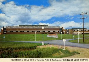 Colour postcard of a large two-storey red brick building on a hill with a sign on front lawn. The signs says Northern College of Applied Arts and Technology - Kirkland Lake Campus.