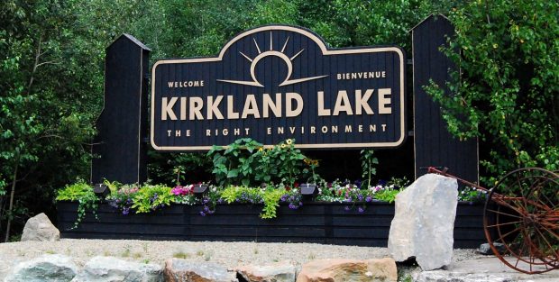 Colour photograph of a large black and gold sign, with the town name of Kirkland Lake in gold lettering. Flowers are planted underneath the sign and large rocks and gravel in front. A piece of mine firefighting equipment is at the right of the picture.