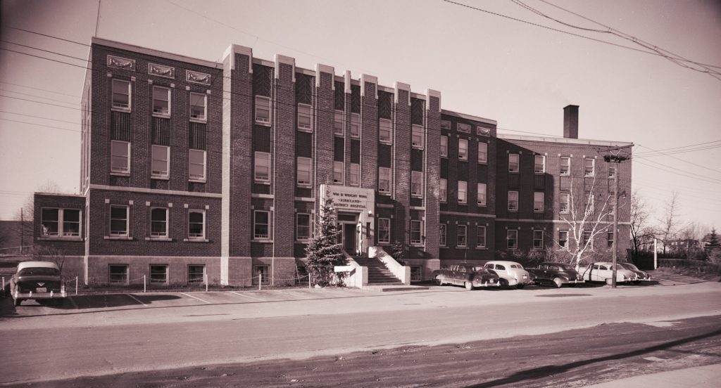 Black and white photograph of the Kirkland District Hospital, a large 4-storey brick building facing onto a street. Cars are parked in front of the building.