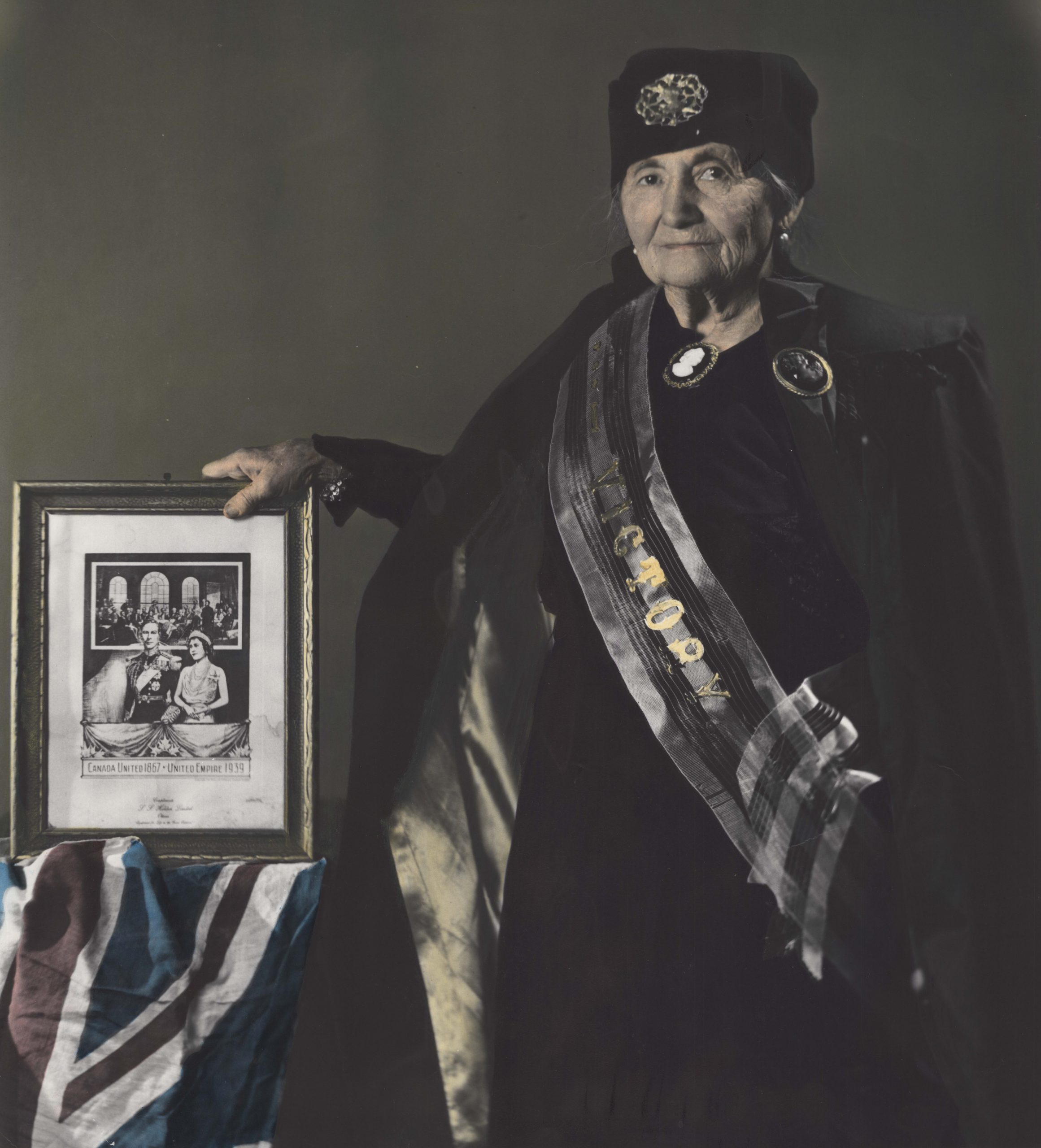 Colourized photograph of Roza Brown as an older woman wearing a dark cape, dress, and hat. A sash with the word Victory in gold lettering is draped over the front of her dress. She is looking at the camera while holding a framed image of King George VI and his wife Queen Elizabeth. A British flag is draped over a table to her left.