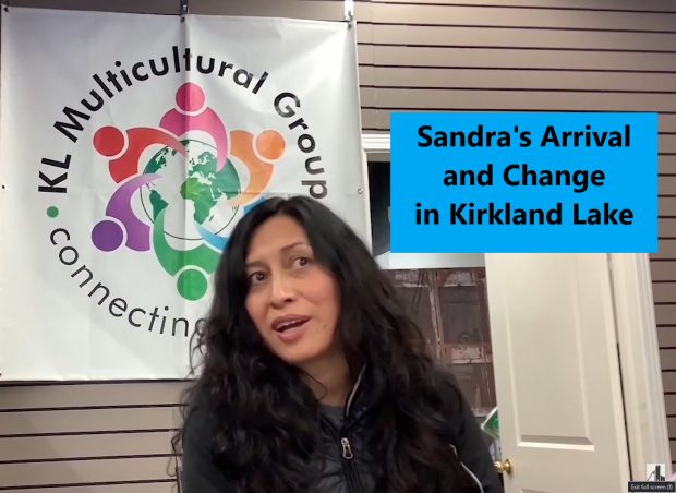 Sandra Reygada Licuime is sitting in front of the Kirkland Lake Multicultural Group sign, facing the camera.