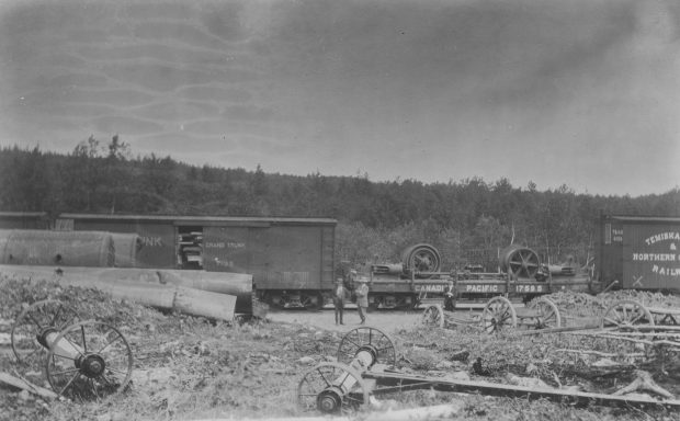 Black and white photograph of large pieces of mining machinery on a railway car. Two men stand in front of the railway car. In the foreground are pieces of metal, large wagons and wheels.