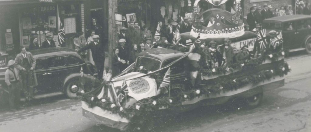 Black and white photograph of a truck in a parade, with people on the sidewalk.