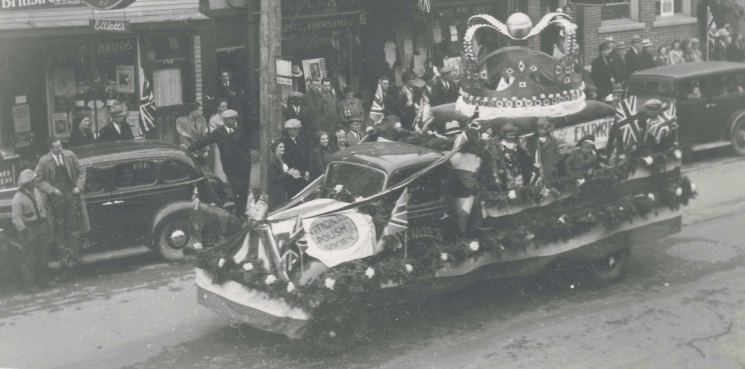 Black and white photograph of a truck in a parade, with people on the sidewalk.