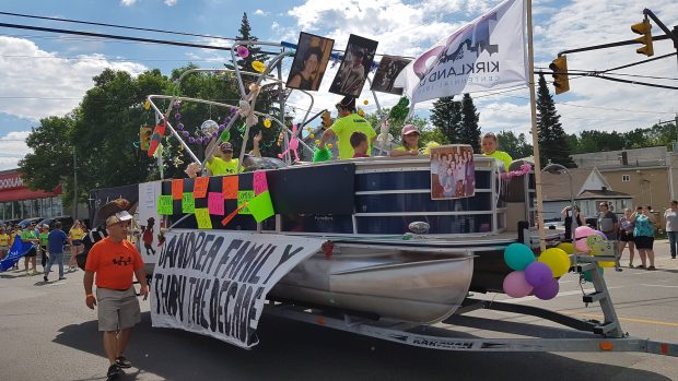 Colour photograph of a large pontoon boat on a trailer is pulled through the street as a float in a parade. The float is decorated with a banner titled D'Andrea Family Thru the Decades hanging off the side with other decorations. At least five people are visible in the float.