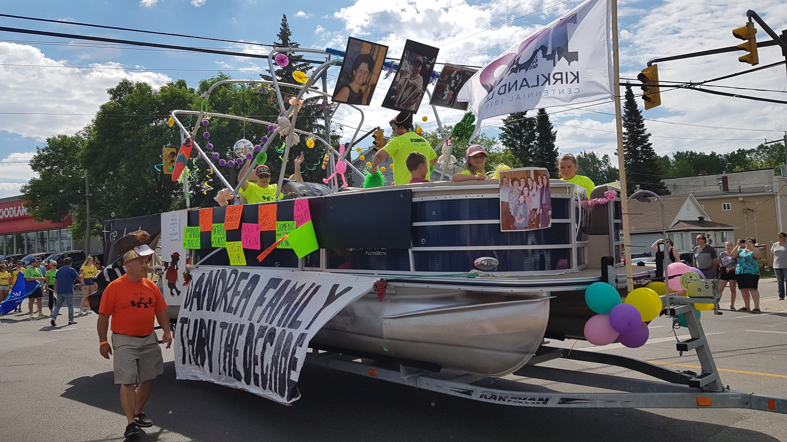 Colour photograph of a large pontoon boat on a trailer is pulled through the street as a float in a parade. The float is decorated with a banner titled D'Andrea Family Thru the Decades hanging off the side with other decorations. At least five people are visible in the float.