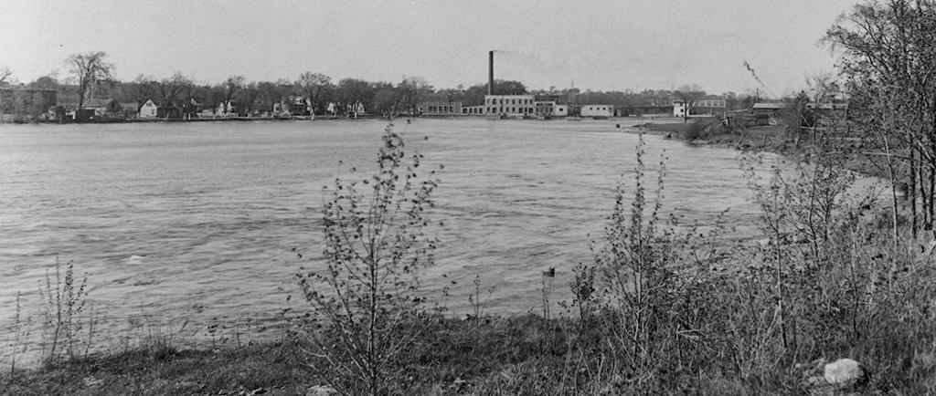 3. Black and white photograph taken by the water basin from behind the Church of the Visitation. To the right on the levee is the Back River Power Company factory with its tall chimney. On the left, the houses on Île-de-la-Visitation Street.