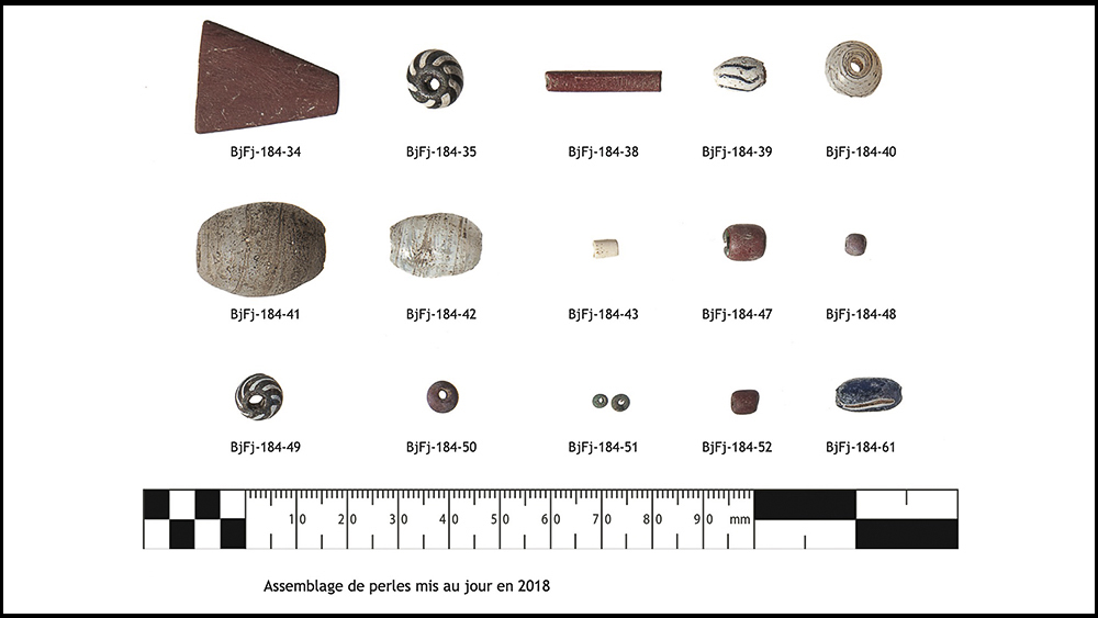 Arrangement of beads found by archaeologists during digs in 2018. It consists of thirteen glass and two red argillite beads. At the bottom of the table, a scale in millimetres.