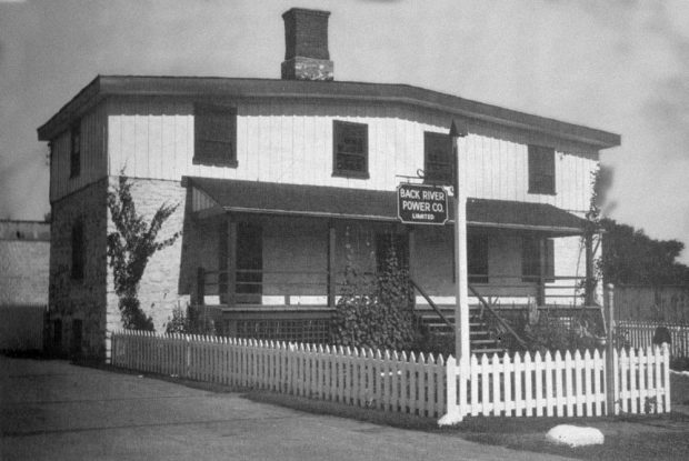 Behind a fence of wooden planks painted white, the building that housed the offices of the Back River Power Company in 1942. Masonry is visible on the ground floor, but there is a different wall colouring on the second floor. A balcony with an inclined roof was added.