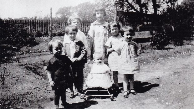 Six of the Danis children huddled around their younger sibling who is in a walker. They are in a garden surrounded by a wooden fence.