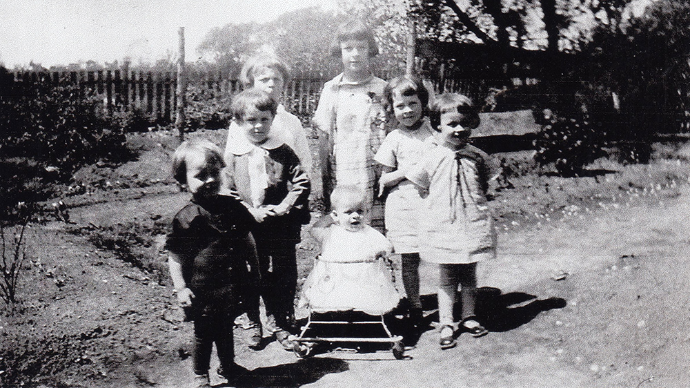 Six of the Danis children huddled around their younger sibling who is in a walker. They are in a garden surrounded by a wooden fence.