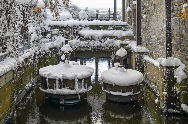 After a snowfall, two hydraulic turbines are covered in white. Today, they lay still above the level of the water going through the dike. To their right, an old masonry wall.