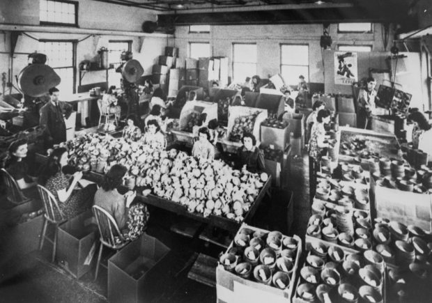 Interior view of the Back River Power Company factory floor in 1942. Women seated at large tables preparing cylindrical cardboard packaging for ammunition that will be shipped to the European front. Standing on the left, a man overseeing their work. On the right, boxes full of cylinders.