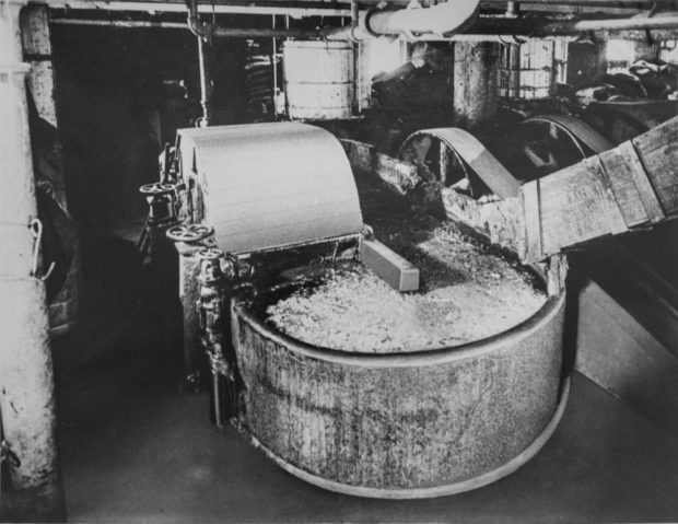 In the centre, a machine is mixing the paper pulp in a circular vat. To the right, big drive belts transfer the torque needed to run large steel-wheeled machines.