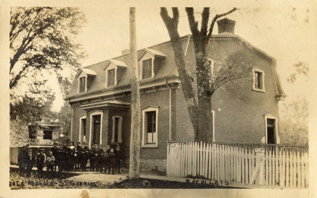 In 1900, the village school, in the centre, looked like a large brick house with a mansard roof and dormer windows. On its left, a group of boys with their teacher. On its right, a wooden fence.