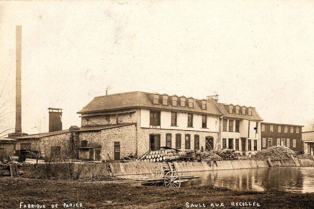 Postcard published around 1900. In the centre: a paper mill, on the left: its tall smokestack overlooking the mill’s dike, cluttered with barrels and other materials. In the foreground: a roll-in dock on the water basin.