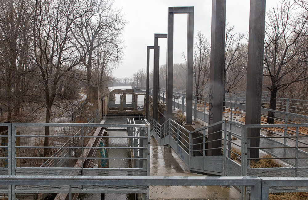 View of the river taken along the belvedere from Du Pont Street on a rainy spring day. On the left, bare trees, on the right, steel structures reminiscent of the volumes of old buildings. In the background, water from the river.