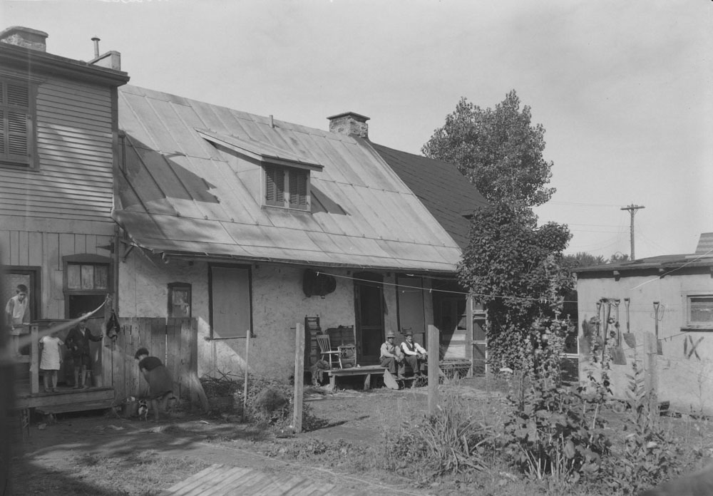 On the left, children playing in the backyard of 10861 Du Pressoir Street. This flat-roofed, two-story home was added to the cider press house. In the centre, two adults are chatting on the back porch of 10865. The cider mill house was divided into two apartments, with differently coloured gable roofs.