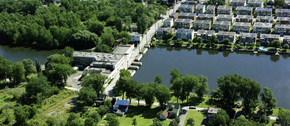 Aerial view of the mills site in 1976. In the centre: the water of the Des Prairies River flows through the dike and abandoned industrial buildings. In the foreground and to the left, the site of the future Île-de-la-Visitation Nature Park. On the right, a row of duplexes.