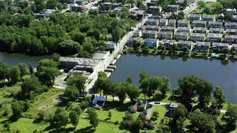 Aerial view of the mills site in 1976. In the centre: the water of the Des Prairies River flows through the dike and abandoned industrial buildings. In the foreground and to the left, the site of the future Île-de-la-Visitation Nature Park. On the right, a row of duplexes.