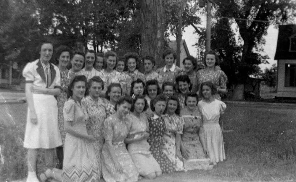 A group of twenty-five female workers split into three rows on De l’Île-de-la-Visitation Street in 1941. They are all wearing different dresses. Behind them, trees and houses on the island.