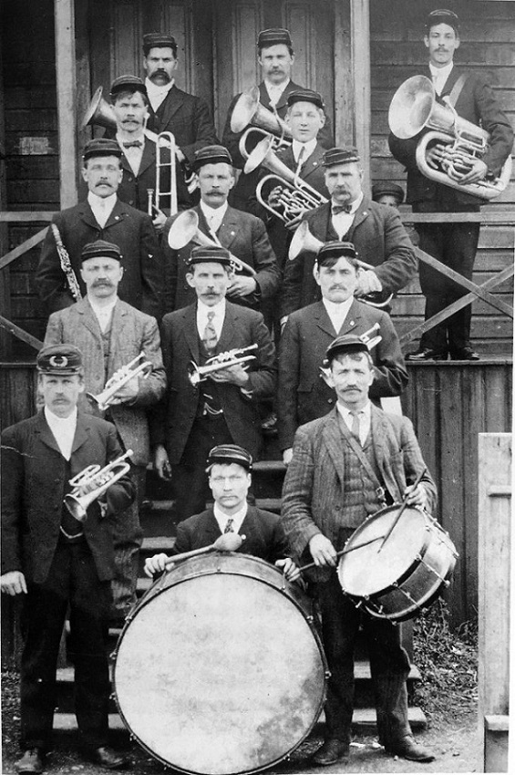 Photo en noir et blanc d'un groupe de musiciens portant des instruments de musique et posant sur les marches d'un bâtiment en bois.