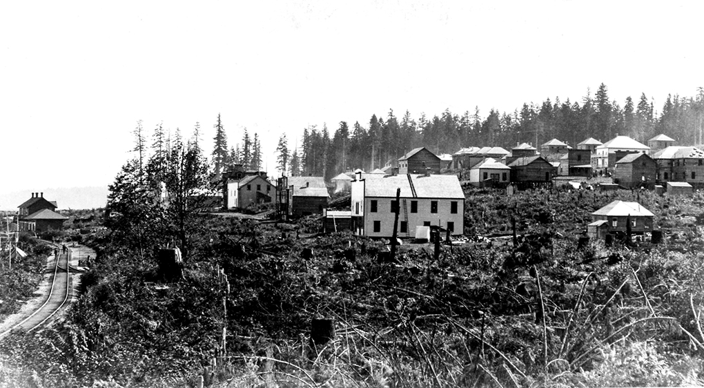 Black and white photograph of a cluster of wooden buildings on a hillside which slopes down toward a set of railway tracks.