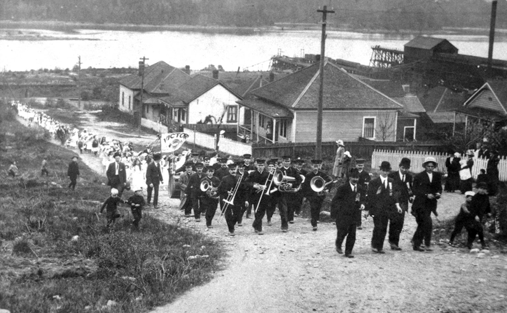 Black and white photograph looking downhill along an unpaved street up which a brass band is leading a procession of women and children. The waters of a harbour form the background.