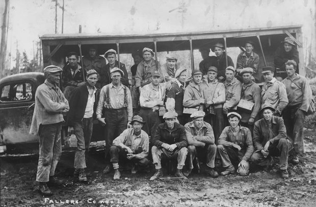 Black and white photograph of a group of 26 men in work clothes, some carrying lunch buckets, posed in front of a truck modified as a crude open-sided bus.