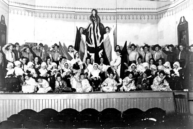 Black and white photograph of a large group of child actors in costume arranged on a stage in three rows, kneeling at the front, sitting in the middle and standing at back. The back row features in its centre three actresses elevated from the rest, each holding flags vertically with a central Union Jack above all.