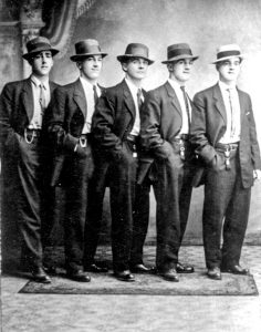 Photographie en noir et blanc de cinq jeunes hommes élégamment vêtus de costumes deux pièces, chemise blanche et cravate avec chapeaux d'époque, debout en échelon à un angle par rapport à la caméra, la main droite dans les poches de leur pantalon.