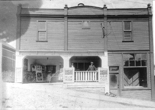 Black and white photograph of a two-storey wooden building on a sloping unpaved street. A large open foyer lined with billboards occupies the uphill two-thirds of the building at ground level. A storefront, apparently empty, occupies the downhill third. Two men stand looking at the photographer.