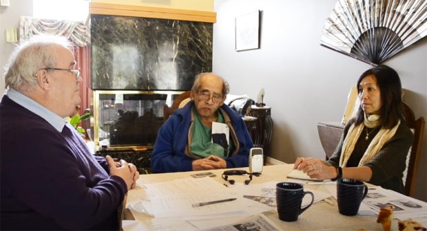 A caucasian male and a Chinese man and woman talking in a dining room. A Chinese wall fan is on the wall behind them.