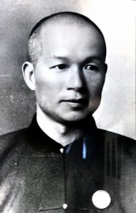Photo d'un portrait tête et épaules en noir et blanc d'un jeune homme chinois aux cheveux coupés très court, rasé de près, portant une veste sans col de couleur sombre de style chinois.