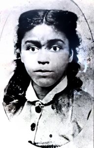 Photo en noir et blanc d'un portrait oval granuleux d'une jeune femme noire aux yeux proéminents et aux longs cheveux noirs bouclés, séparés au milieu et ramenés derrière le visage.