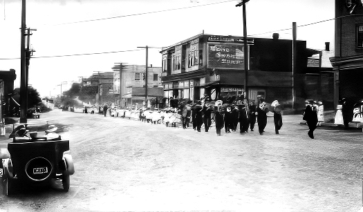 Photo d'époque en noir et blanc d'une rue non pavée bordée de bâtiments commerciaux de deux et trois étages, le long de laquelle un défilé est mené par une fanfare en uniforme composée d'environ 15 musiciens, suivie de plus d'une centaine d'enfants, dont beaucoup sont vêtus de blanc et coiffés de chapeaux blancs, marchant en rangs de trois. Ils sont suivis par une file de véhicules qui s'étend au-delà du champ de vision.