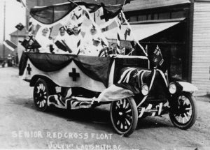 Photo en noir et blanc, vue oblique, d’un camion à roues à rayons à caisse ouverte et à long châssis, orné de drapeaux Union Jack et Canada Red Ensign sur le capot et les marchepieds. Le conducteur, vêtu de blanc et coiffé d’une casquette blanche, est assis au volant. Derrière lui, la caisse, dont les côtés sont décorés de croix en tissu, contient au moins cinq infirmières assises, vêtues d’uniformes blancs et coiffées de foulards blancs.