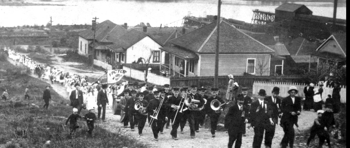 Black and white photograph looking downhill along an unpaved street up which a brass band is leading a procession of women and children. The waters of a harbour form the background.