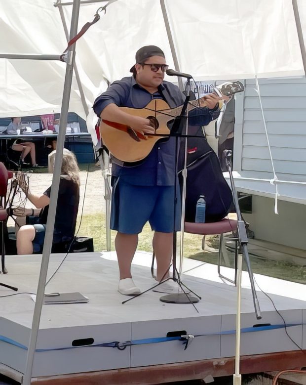 Colour photograph of a Coast Salish musician standing on a stage in front of a microphone playing an acoustic electric guitar and singing.