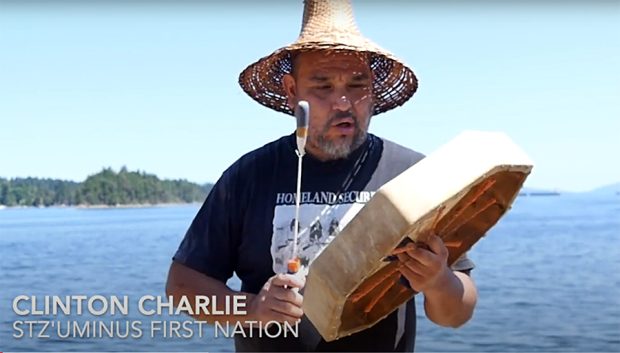Colour photo of Coast Salish man wearing a traditional Cedar hat, and drumming a ceremonial drum.