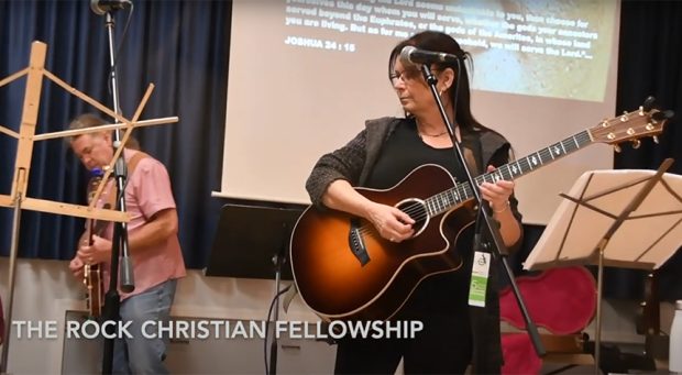 Colour photo of two performers with guitars on a stage. Slide show behind them with Scripture.