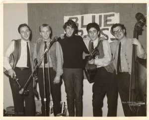 Photographie en noir et blanc de quatre hommes portant des gilets et des cravates et tenant des guitares, des mandolines et une basse sur pied, avec au centre un cinquième homme portant un pull à col roulé noir.