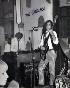 Black and white photograph of a man with a black vest, checkered pants and long hair playing guitar on a small stage. Behind him, a man with a beret and sunglasses is playing the organ. Spectators are seen crowded along the left side of the image.