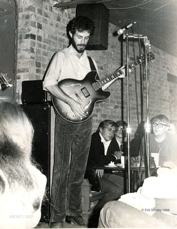 Black and white photo of singer Jesse Winchester with medium length black hair, moustache and trimmed beard holding guitar at a café with patrons seated next to the stage.