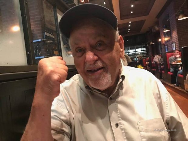 Close up photo of a white-haired elderly Larry Kent seated at a table in a restaurant wearing a cap and with a clenched fist held up towards the camera.