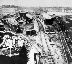 Wagons alignés d’un côté de la gare de triage de la Great Western Railway. On voit aussi sur l’image des piles de bois et d’autres matériaux sur les voies. Deux travailleurs se trouvent à l’avant-plan.