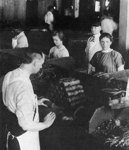 Young women and children working on the shop floor of Tuckett and Billings Tobacco Manufactory. The women are dressed in 19th century uniforms, with many wearing white work aprons. Rolls of tobacco sit in front of them and one young woman looks directly at the camera.
